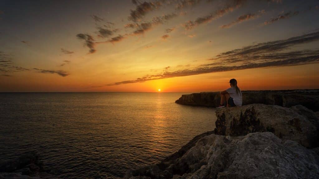 Person sitting alone on a cliff watching the sunset over the ocean, representing calm reflection and anxiety relief through therapy