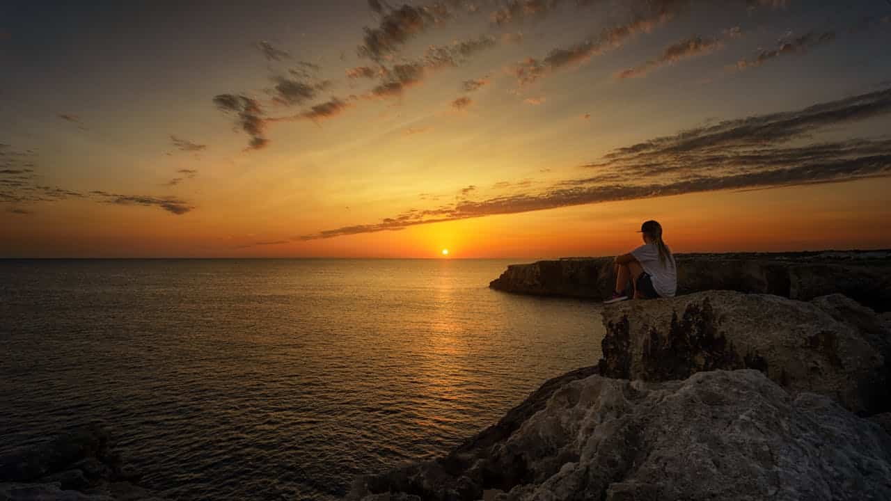 Person sitting alone on a cliff watching the sunset over the ocean, representing calm reflection and anxiety relief through therapy